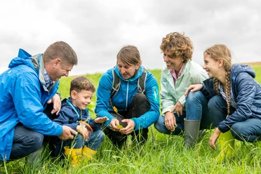 Natur entdecken mit den Naturentdeckern aus Wülfrath Familiengruppe erkundet gemeinsam eine grüne Wiese, alle lächeln und tragen wetterfeste Kleidung.
