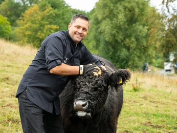 Dirk Hanten und Galloway-Rind Dirk Hanten streichelt ein Galloway-Rind auf einer grünen Wiese, im Hintergrund sind Bäume.