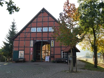Haus des Gastes Mardorf Backsteingemäuer im Fachwerkstil mit Baum im Vordergrund, geschützt durch Bäume und Infotafeln flankiert.Brick walls in half-timbered style with a tree in the foreground, protected by trees and flanked by information boards.Murstensvægge i bindingsværksstil med et træ i forgrunden, beskyttet af træer og flankeret af informationstavler.Bakstenen muren in vakwerkstijl met een boom op de voorgrond, beschermd door bomen en geflankeerd door informatieborden.