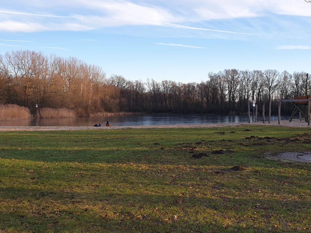 Ein ruhiger Badesee mit Sandstrand und Spielplatz, umgeben von Bäumen und grasbewachsenen Ufern.