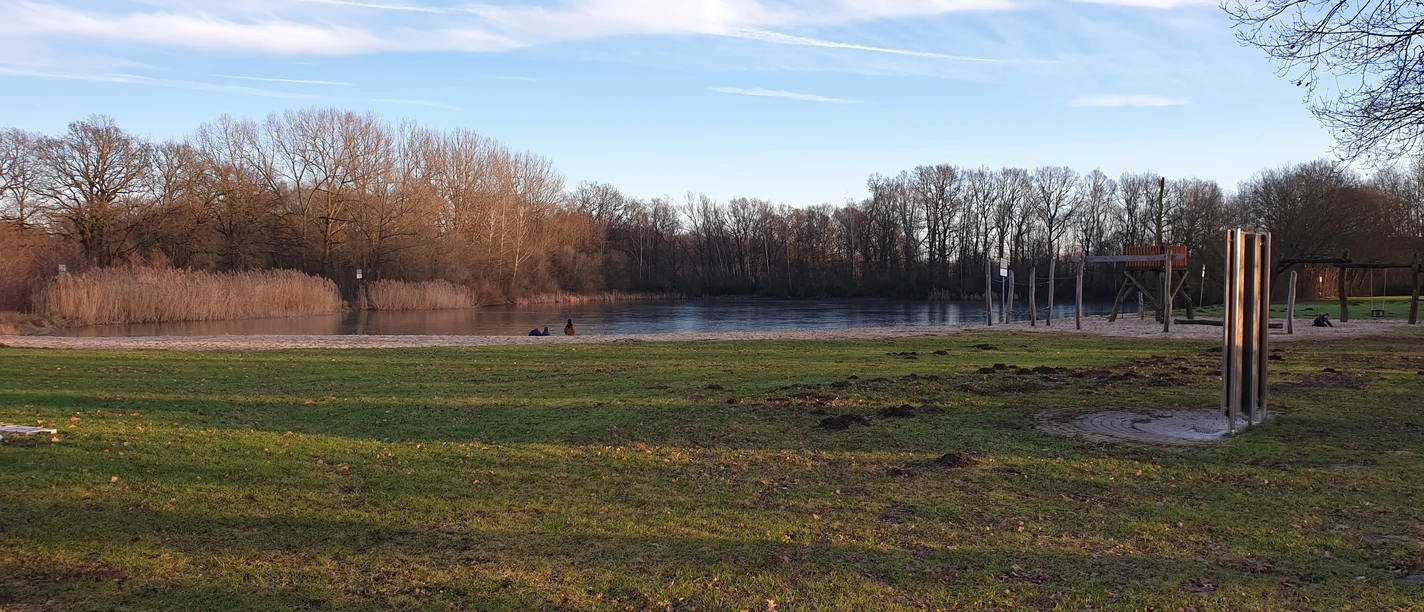Badesee Mindenerwald Ein ruhiger Badesee mit Sandstrand und Spielplatz, umgeben von Bäumen und grasbewachsenen Ufern.