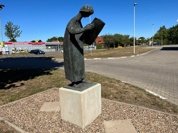 A metal sculpture depicts a figure curiously leaning over a newspaper against a blue sky.
