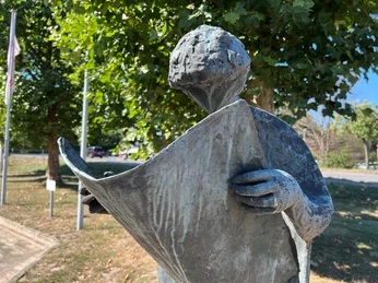 Bronzeskulptur eines Menschen, der vertieft in eine Zeitung liest, umgeben von grünen Bäumen.