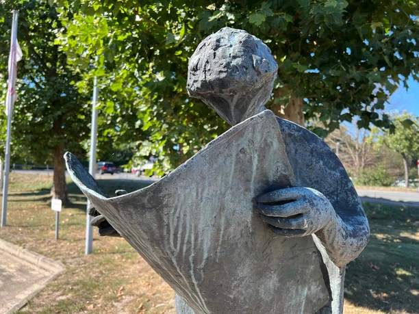 Bronzeskulptur eines Menschen, der vertieft in eine Zeitung liest, umgeben von grünen Bäumen.