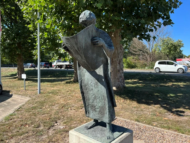 Bronzestatue eines Mannes liest stehend eine offene Zeitung neben Straße und grünen Bäumen.