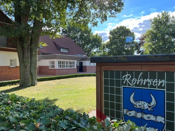 Gebäude mit Ziegeldach im Grünen, nahe Schild mit Ortsnamen "Rohrsen" und einem Wappen im Vordergrund.