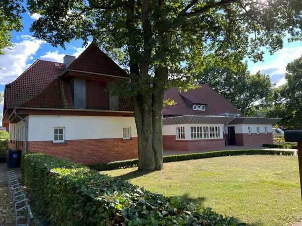 Alte Schule Rohrsen Ein Backsteingebäude mit rotem Dach und großen Bäumen, umgeben von Grünflächen und Hecken.