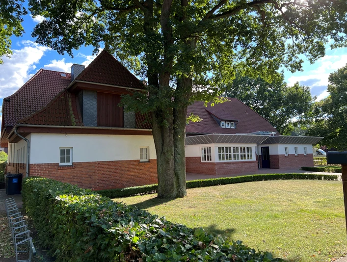 Alte Schule Rohrsen Ein Backsteingebäude mit rotem Dach und großen Bäumen, umgeben von Grünflächen und Hecken.