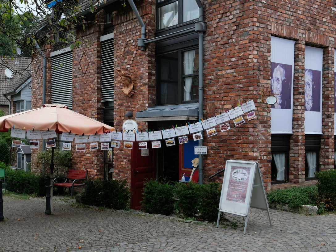 Cafe Im Dorf in Haan-Gruiten Ein Café mit roter Backsteinfassade, schattenspendendem Sonnenschirm und dekorativen Girlanden.