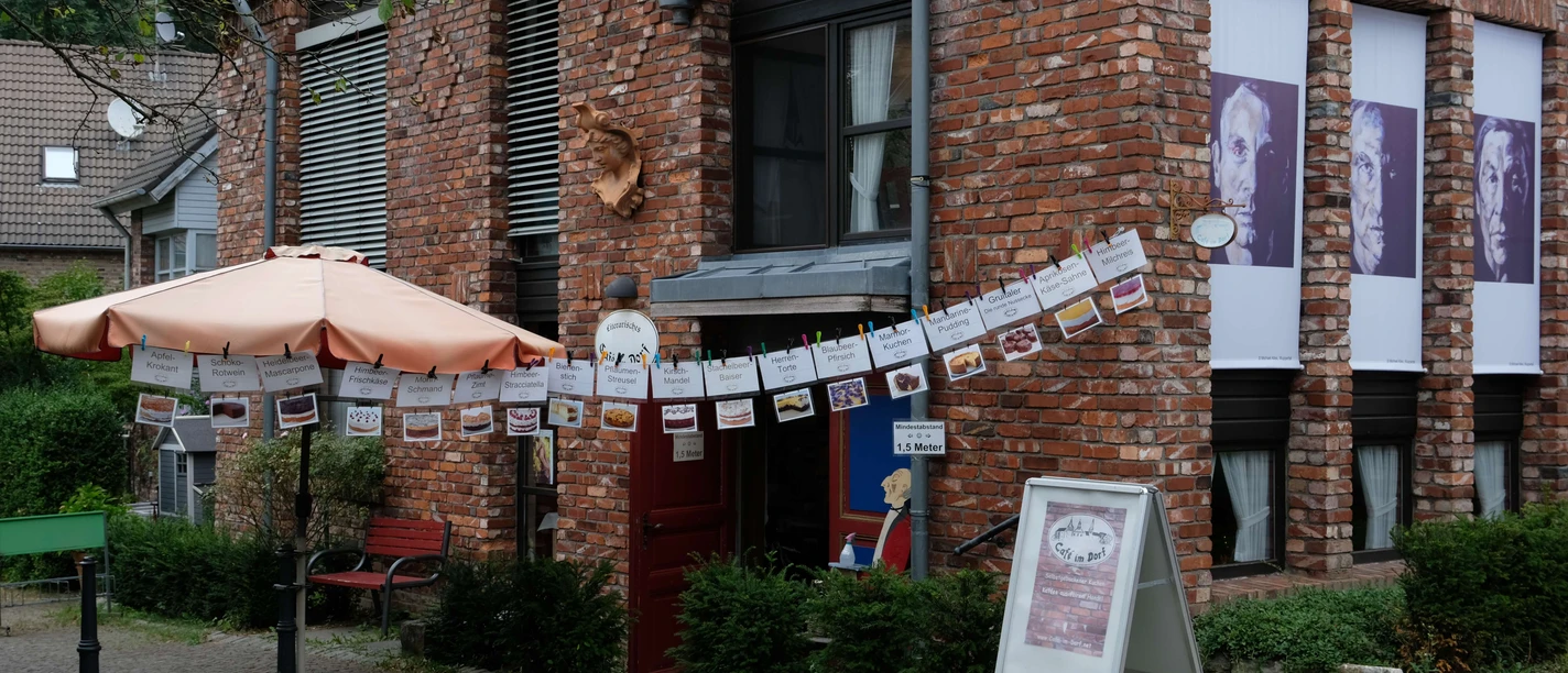 Cafe Im Dorf in Haan-Gruiten Ein Café mit roter Backsteinfassade, schattenspendendem Sonnenschirm und dekorativen Girlanden.