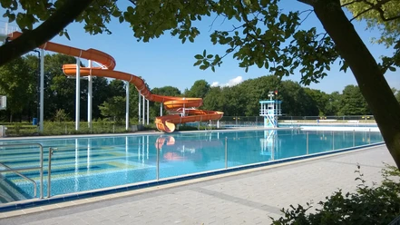 Stadtbad Langenfeld Orange Wasserrutsche im Freibad Langenfeld neben einem klaren Becken, umgeben von Bäumen und Gelände.