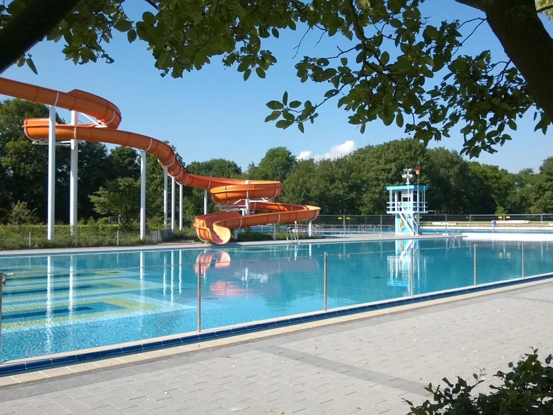 Stadtbad Langenfeld Orange Wasserrutsche im Freibad Langenfeld neben einem klaren Becken, umgeben von Bäumen und Gelände.