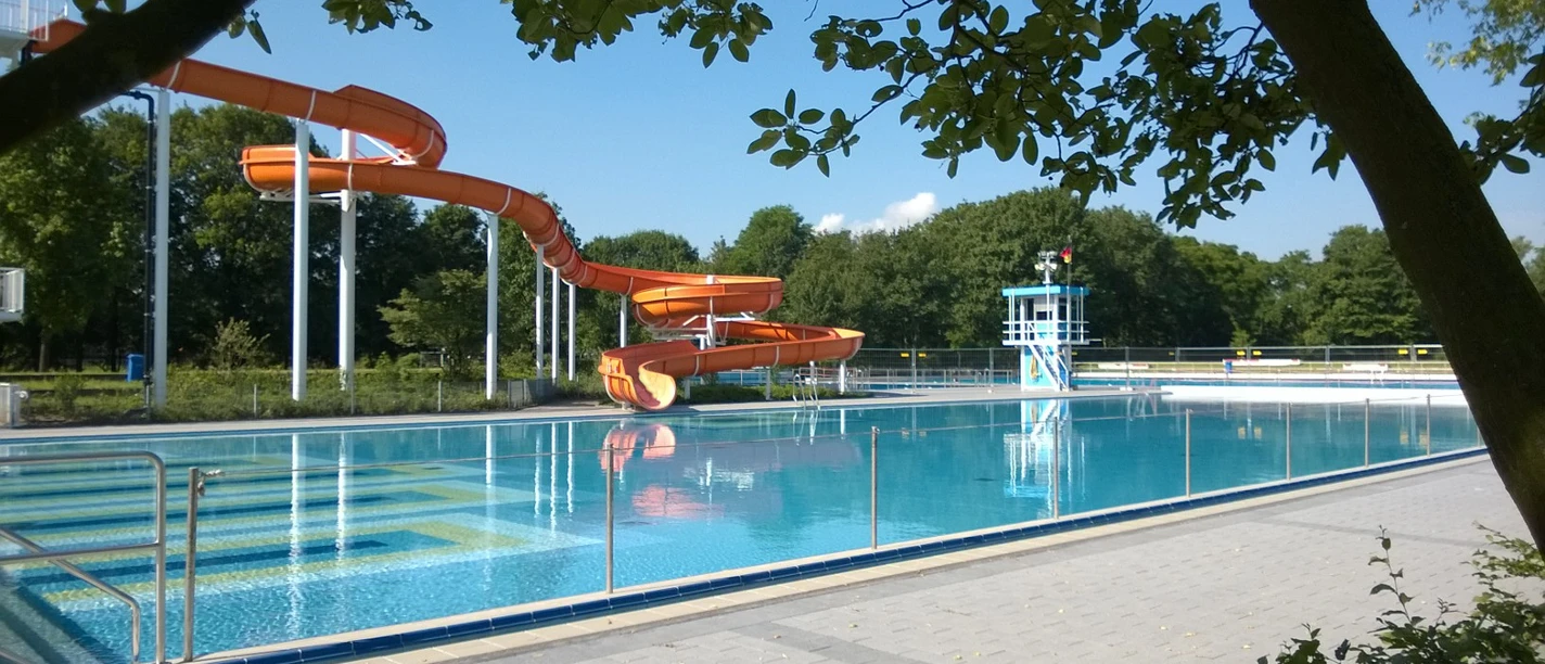 Stadtbad Langenfeld Orange Wasserrutsche im Freibad Langenfeld neben einem klaren Becken, umgeben von Bäumen und Gelände.