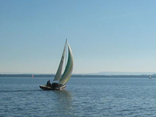 Wassersport Segelboot mit buntem Segel gleitet bei blauem Himmel über einen ruhigen, weitläufigen See.