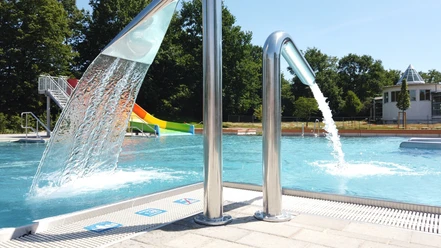 Ein Freibad mit Wasserspielen und Rutschen im Hintergrund, umgeben von Bäumen und blauen Himmel.