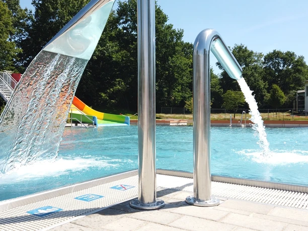 Ein Freibad mit Wasserspielen und Rutschen im Hintergrund, umgeben von Bäumen und blauen Himmel.