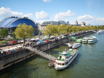 Köln-Düsseldorfer Deutsche Rheinschiffahrt Ein Schiffsanleger am Rhein mit mehreren anliegenden Passagierschiffen, umgeben von Bäumen und einer Promenade.A jetty on the Rhine with several adjacent passenger ships, surrounded by trees and a promenade.