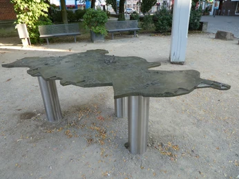 Metallmodell der Stadt Achim auf stabilen Säulen, umgeben von Bänken und Bäumen auf einem Platz.