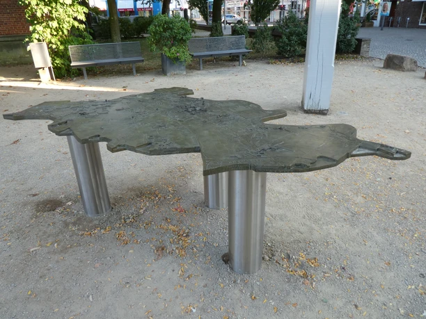 A metal model of the city of Achim on sturdy pillars, surrounded by benches and trees in a square.