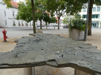 Bronzenes Stadtmodell von Achim auf belebtem Platz, umgeben von Bäumen und modernen Gebäuden.