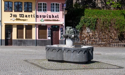Fischweiberbrunnen Vor einem historischen Brauhaus steht der Fischbrunnen mit einem steinernen Becken und einer Figurengruppe als zentralem Element. Die Fassaden der umliegenden Gebäude zeigen sanfte Pastelltöne, die eine einladende Atmosphäre schaffen.