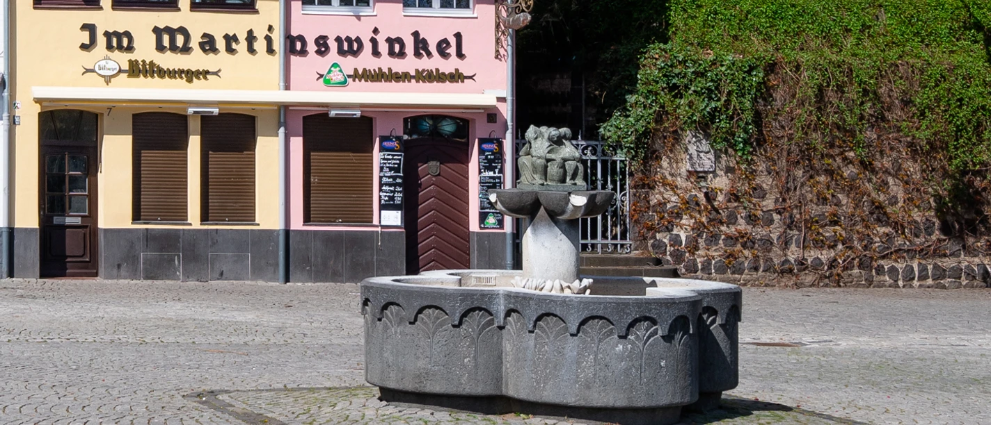 Fischweiberbrunnen Vor einem historischen Brauhaus steht der Fischbrunnen mit einem steinernen Becken und einer Figurengruppe als zentralem Element. Die Fassaden der umliegenden Gebäude zeigen sanfte Pastelltöne, die eine einladende Atmosphäre schaffen.
