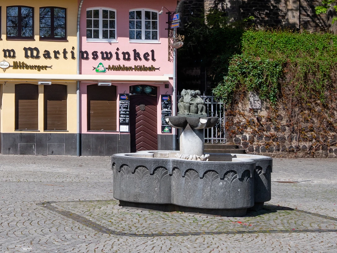 Fishwife fountain Vor einem historischen Brauhaus steht der Fischbrunnen mit einem steinernen Becken und einer Figurengruppe als zentralem Element. Die Fassaden der umliegenden Gebäude zeigen sanfte Pastelltöne, die eine einladende Atmosphäre schaffen.The fish fountain with a stone basin and a group of figures as its central element stands in front of a historic brewery. The façades of the surrounding buildings feature soft pastel shades that create an inviting atmosphere.