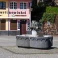 Fishwife fountain Vor einem historischen Brauhaus steht der Fischbrunnen mit einem steinernen Becken und einer Figurengruppe als zentralem Element. Die Fassaden der umliegenden Gebäude zeigen sanfte Pastelltöne, die eine einladende Atmosphäre schaffen.The fish fountain with a stone basin and a group of figures as its central element stands in front of a historic brewery. The façades of the surrounding buildings feature soft pastel shades that create an inviting atmosphere.
