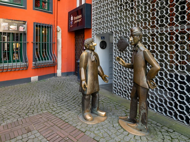 Tünnes and Schäl Monument The bronze figures of Tünnes and Schäl stand on a cobbled path in front of a bright red façade. The figures appear to be engrossed in a lively conversation.