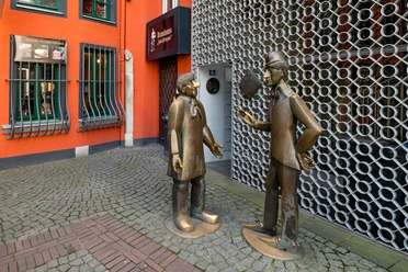 Tünnes-und-Schäl-Denkmal Die Bronzefiguren von Tünnes und Schäl stehen auf einem gepflasterten Weg vor einer leuchtend roten Fassade. Die Figuren scheinen in ein lebhaftes Gespräch vertieft zu sein.