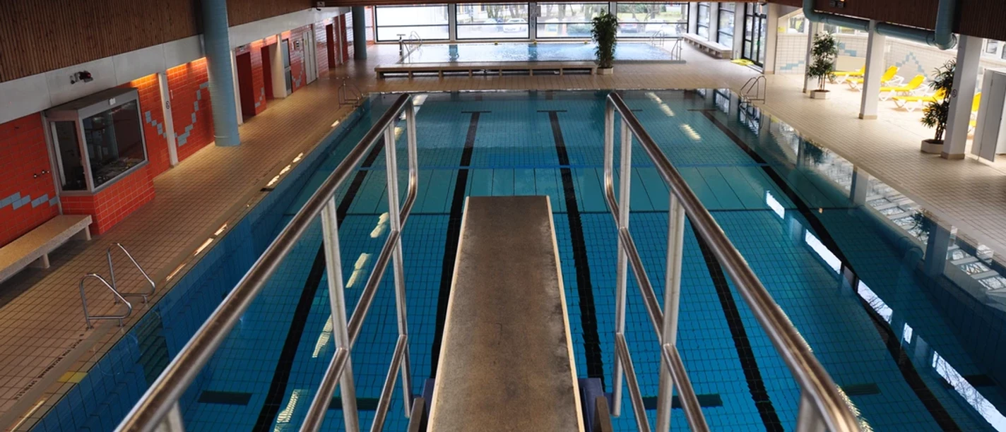 Blick auf ein Hallenbad mit Sprungturm, hellem Raum, großen Fenstern und einem Schwimmbecken.