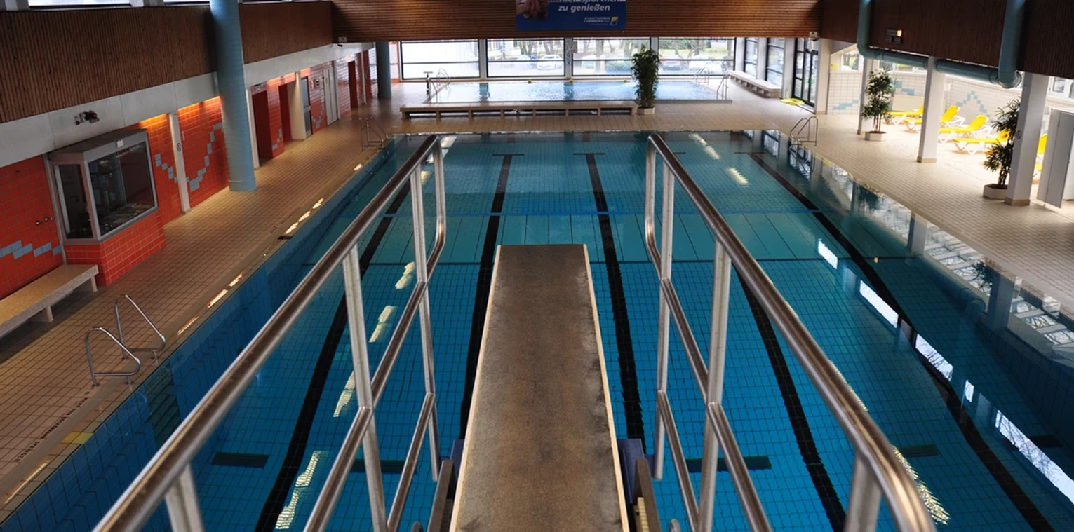 Blick auf ein Hallenbad mit Sprungturm, hellem Raum, großen Fenstern und einem Schwimmbecken.