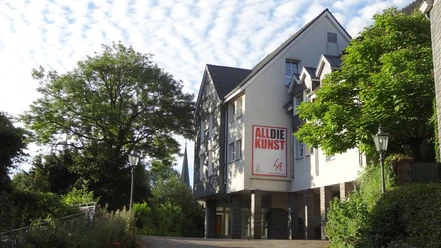 ALLDIEKUNST im Kunsthaus Velbert-Langenberg Ein Kunstzentrum in hellem Gebäude, umgeben von grünen Bäumen und einem klaren blauen Himmel.