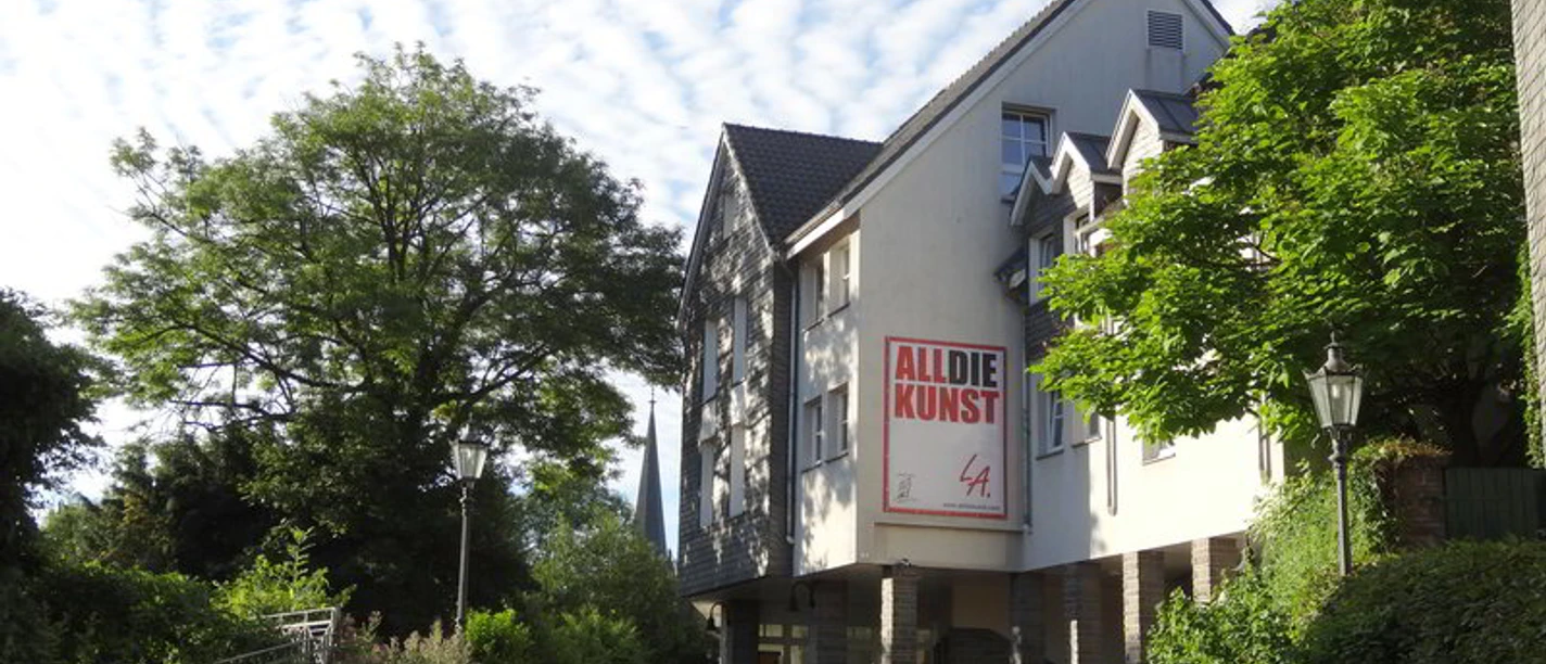 ALLDIEKUNST im Kunsthaus Velbert-Langenberg Ein Kunstzentrum in hellem Gebäude, umgeben von grünen Bäumen und einem klaren blauen Himmel.