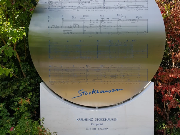 Stockhausen Grabsstätte Gedenkstein von Karlheinz Stockhausen mit Notenblatt, umgeben von üppigem Grün und weißen Steinen.