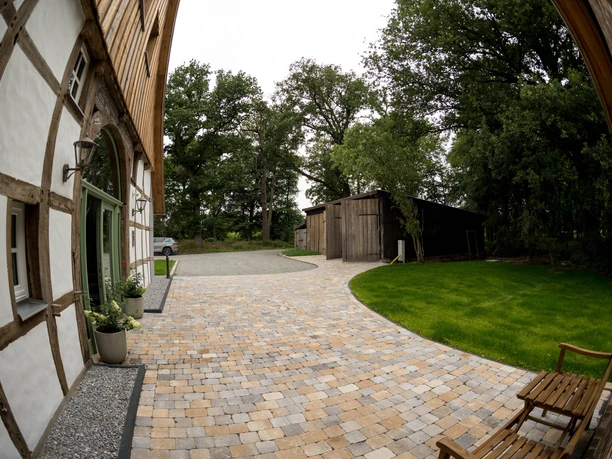 heuerling-2928.jpg Hofeingang mit Fachwerkhaus, gepflasterter Weg zu Scheune, umgeben von Bäumen und gepflegtem Rasen.