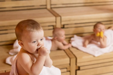 SWO_BabySauna_132.jpg Three babies relax on wooden benches in a sauna; one chews on a towel, two rest behind it.