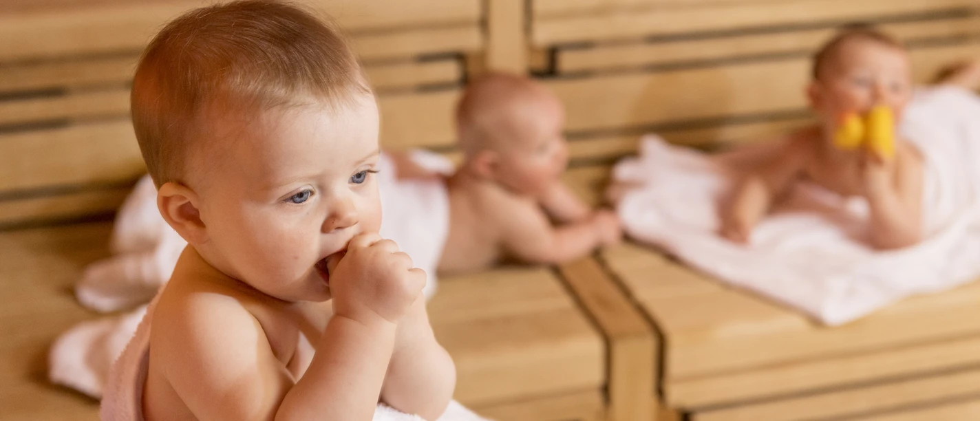 SWO_BabySauna_132.jpg Drei Babys entspannen auf Holzbänken in einer Sauna; eines kaut auf einem Handtuch, zwei ruhen dahinter.
