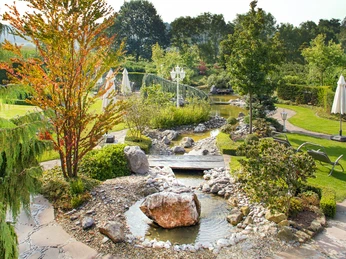 Ringhotel Teutoburger Wald_Gästegarten 2_ Quelle Ringhotel Teutoburger Wald.jpg Gepflegter Hotelgarten mit Steinen, Bachlauf und Liegestühlen in grüner, bewaldeter Umgebung.