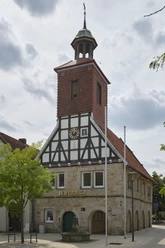 1-SMT-Wehnert_DSCF3450-1.jpg Historisches Fachwerkgebäude mit markantem Uhrturm und steinernen Rundbögen vor bewölktem Himmel.