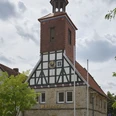 1-SMT-Wehnert_DSCF3450-1.jpg Historisches Fachwerkgebäude mit markantem Uhrturm und steinernen Rundbögen vor bewölktem Himmel.