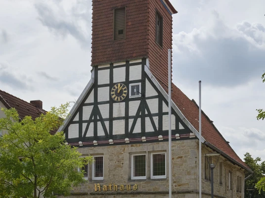 1-SMT-Wehnert_DSCF3450-1.jpg Historisches Fachwerkgebäude mit markantem Uhrturm und steinernen Rundbögen vor bewölktem Himmel.