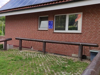 Behinderten-Parkplatz bei einem roten Backsteingebäude mit einem Holzgeländer im Vordergrund.