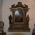 Holzaltar mit Barockmalerei, flankiert von Heiligenstatuen. Zwei gestreifte Stühle stehen daneben.
