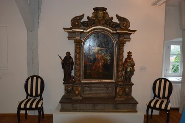 DSC_6985.JPG Holzaltar mit Barockmalerei, flankiert von Heiligenstatuen. Zwei gestreifte Stühle stehen daneben.