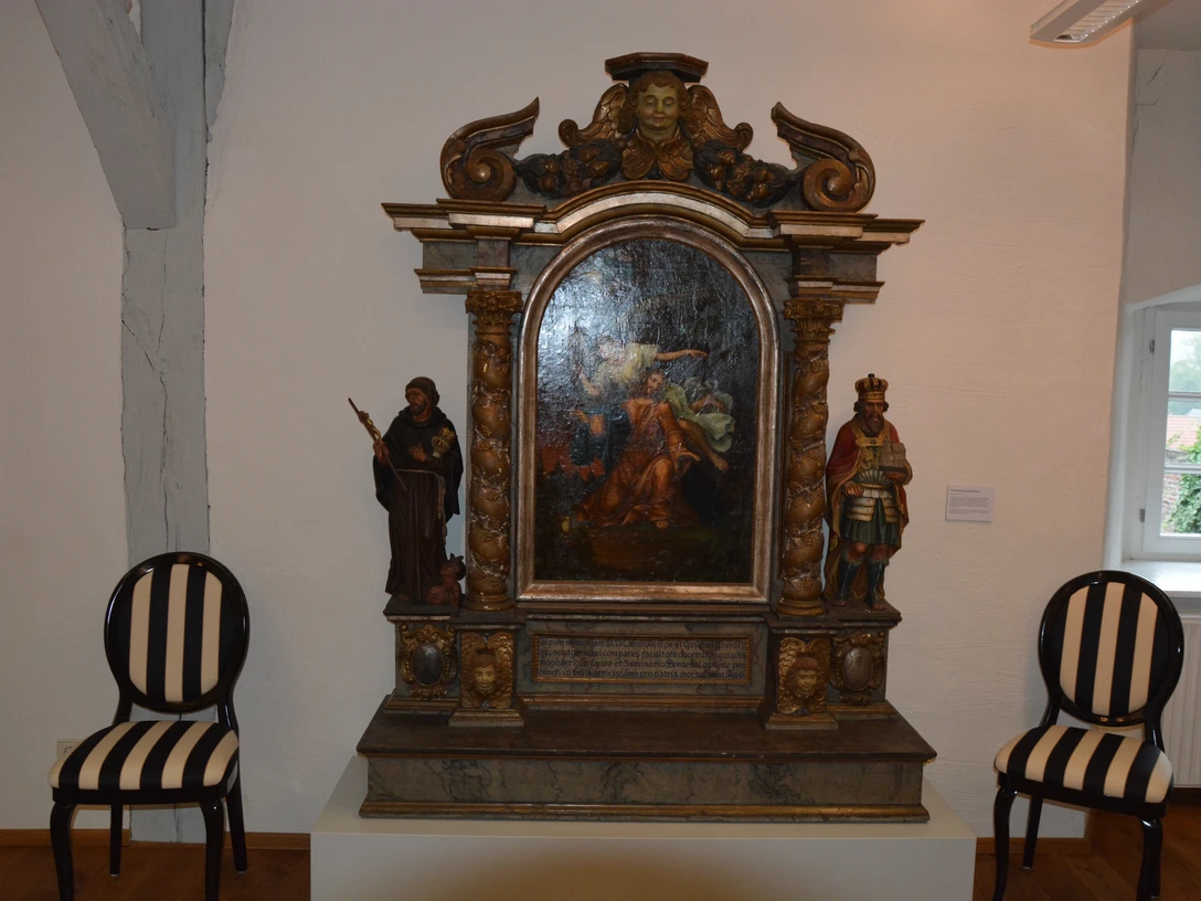 DSC_6985.JPG Holzaltar mit Barockmalerei, flankiert von Heiligenstatuen. Zwei gestreifte Stühle stehen daneben.