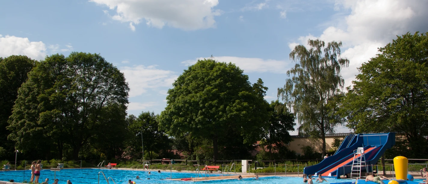 Ein Freibad mit großer Wasserrutsche, umgeben von Bäumen unter einem blauen Himmel mit Wolken.
