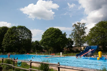 Freibad Lahde Ein Freibad mit großer Wasserrutsche, umgeben von Bäumen unter einem blauen Himmel mit Wolken.