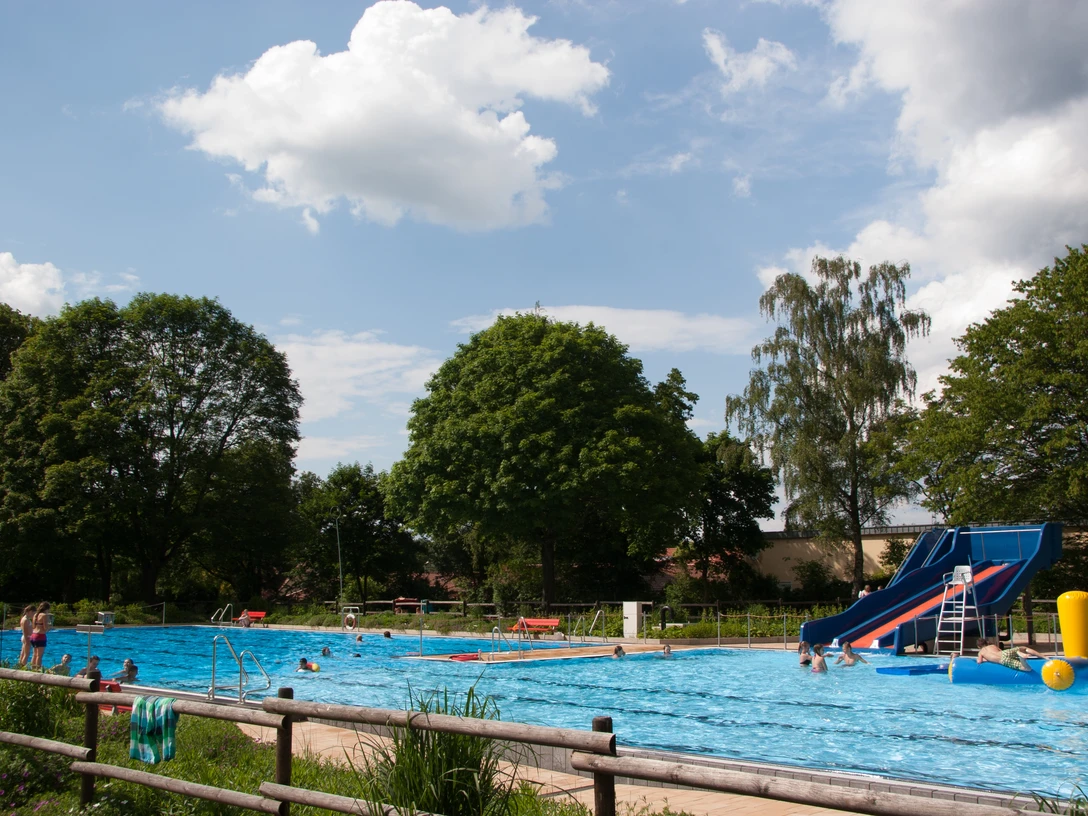 Freibad Lahde Ein Freibad mit großer Wasserrutsche, umgeben von Bäumen unter einem blauen Himmel mit Wolken.