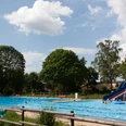Freibad Lahde Ein Freibad mit großer Wasserrutsche, umgeben von Bäumen unter einem blauen Himmel mit Wolken.